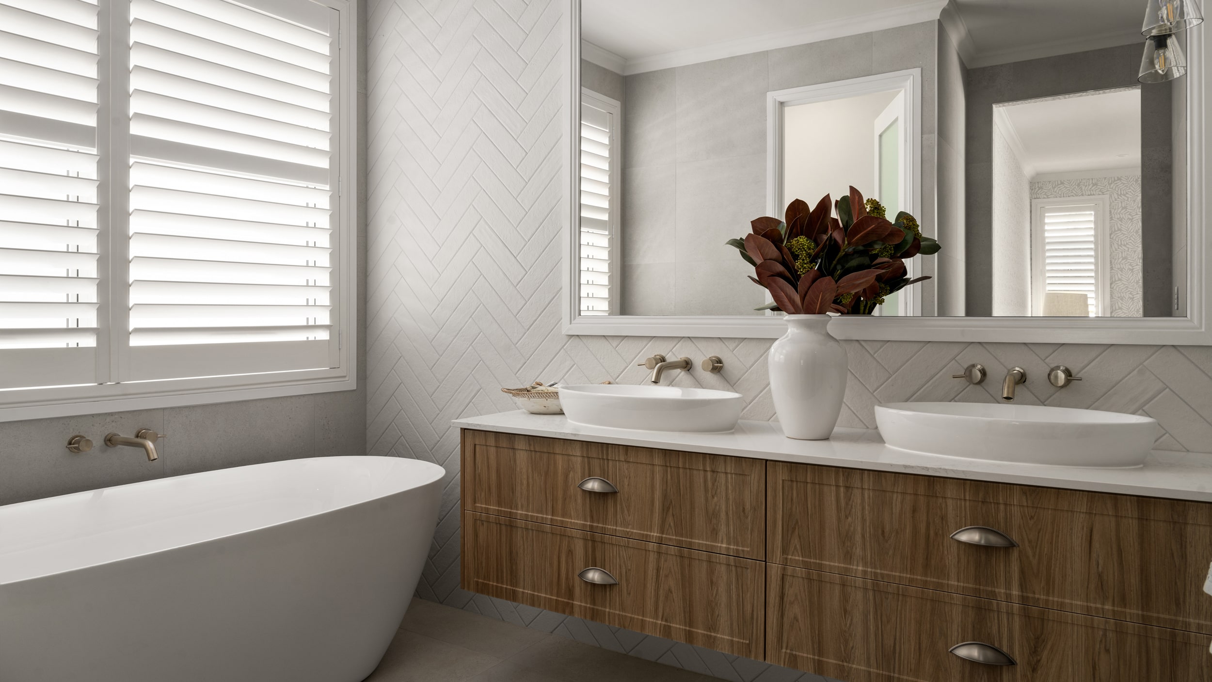A stylish bathroom featuring wide timber vanity, two white basins and large mirror, next to a deep soaking bath.