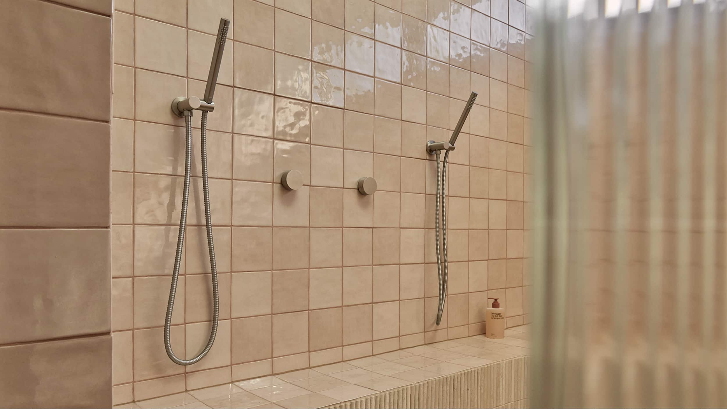 An expansive shower room, featuring blush tile, two microphone-style handshowers, long bench seat and fluted glass shower screen
