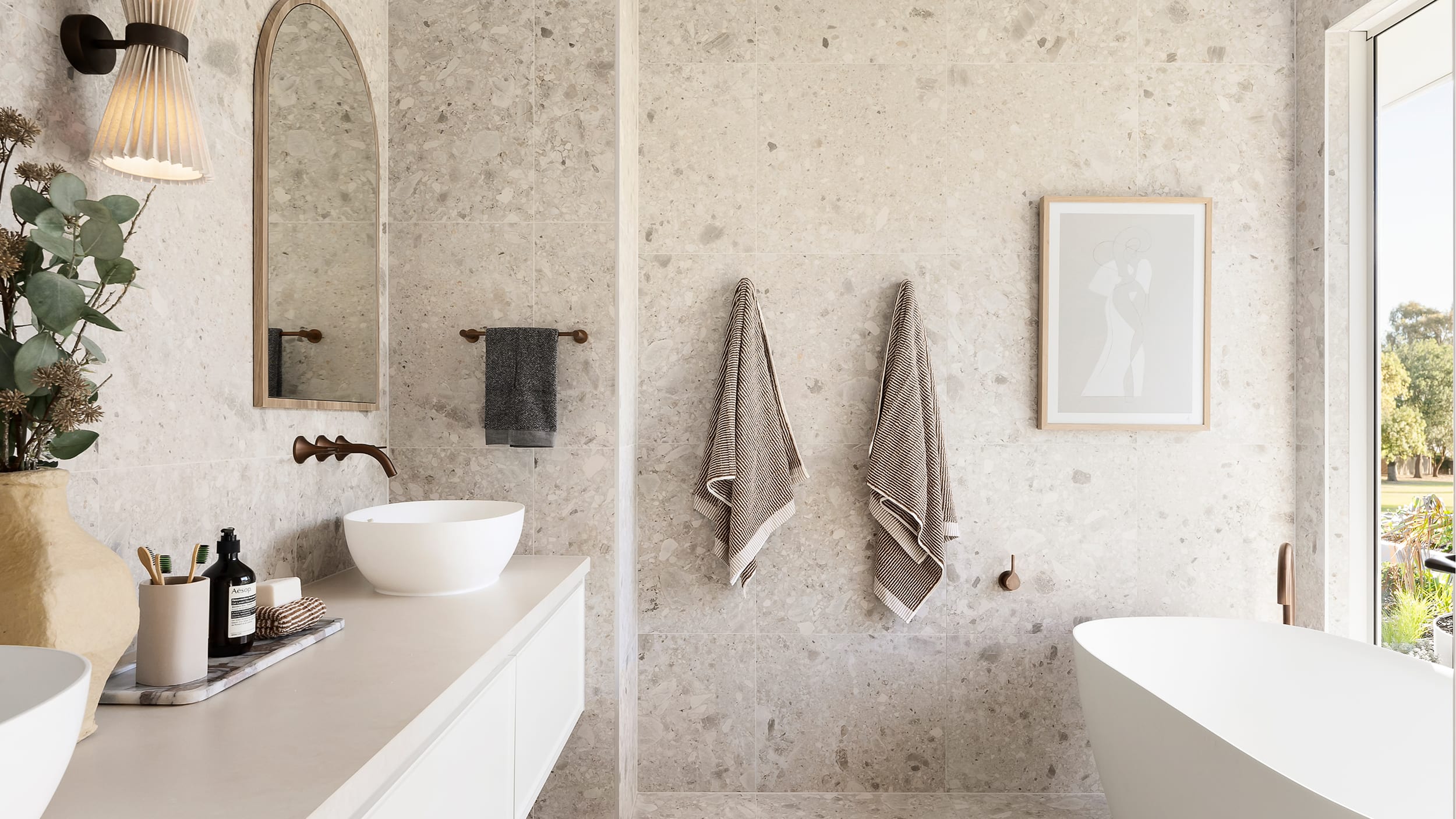 A light-filled, spacious bathroom with a large, full-length window on one side, tiled with large, light grey tiles, furnished with a clean white freestanding bathtub, light vanity and dark, brushed metal tapware.
