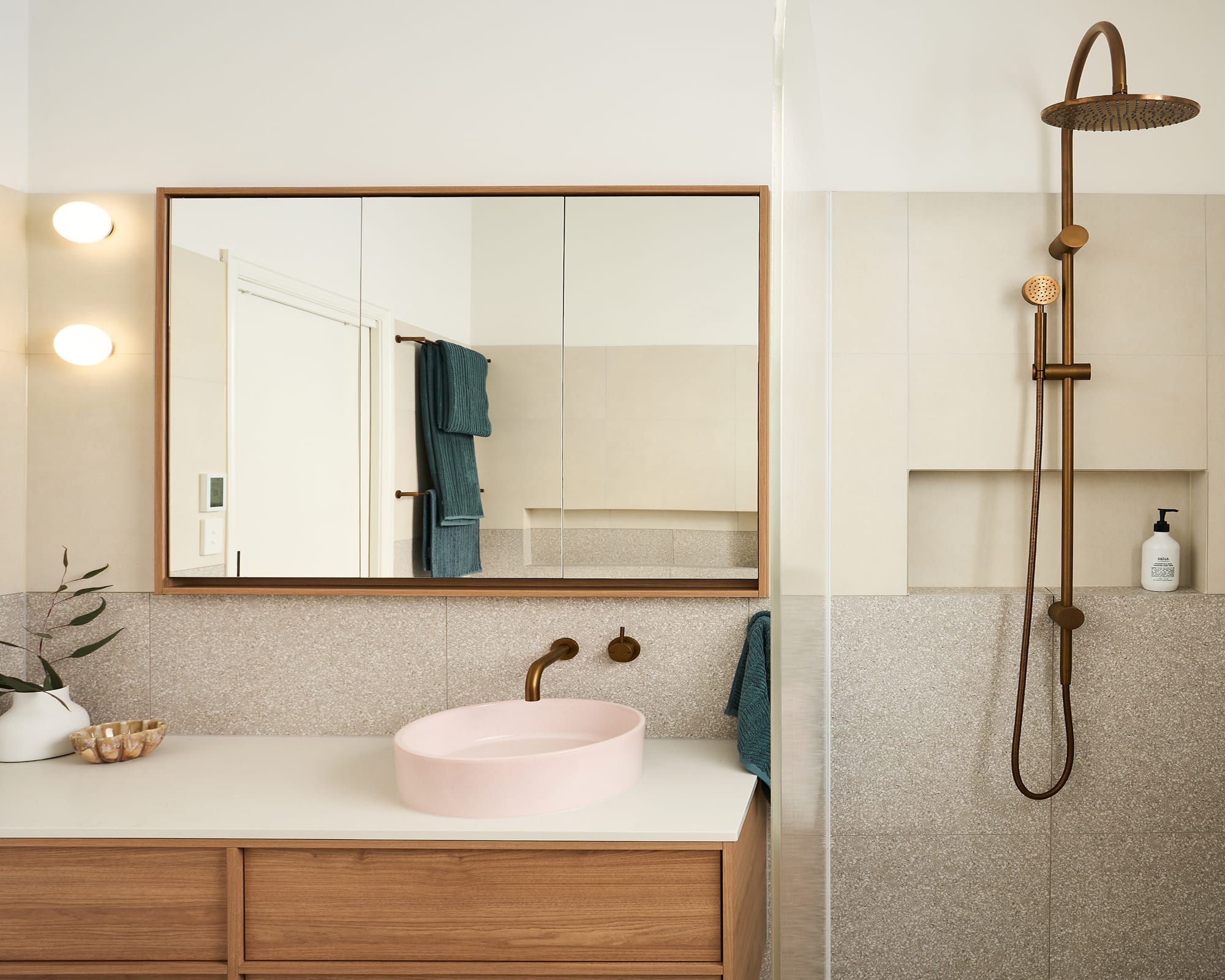 A sleek, stylish bathroom featuring a pink basin, wooden vanity, and dark brushed metal tapware, blending colour and natural materials.