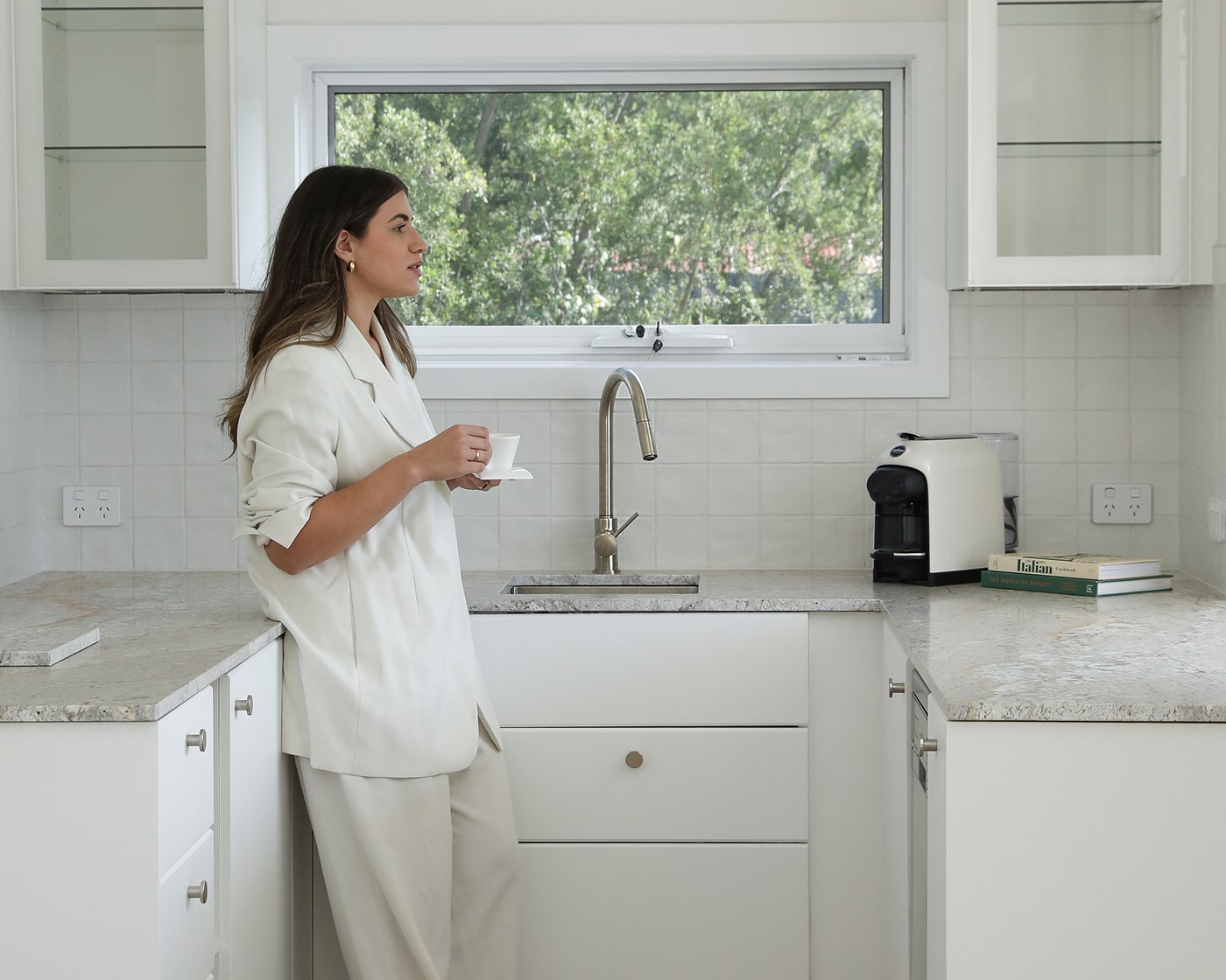 A woman holding a cup of tea, standing relaxed, next to a sink and window.