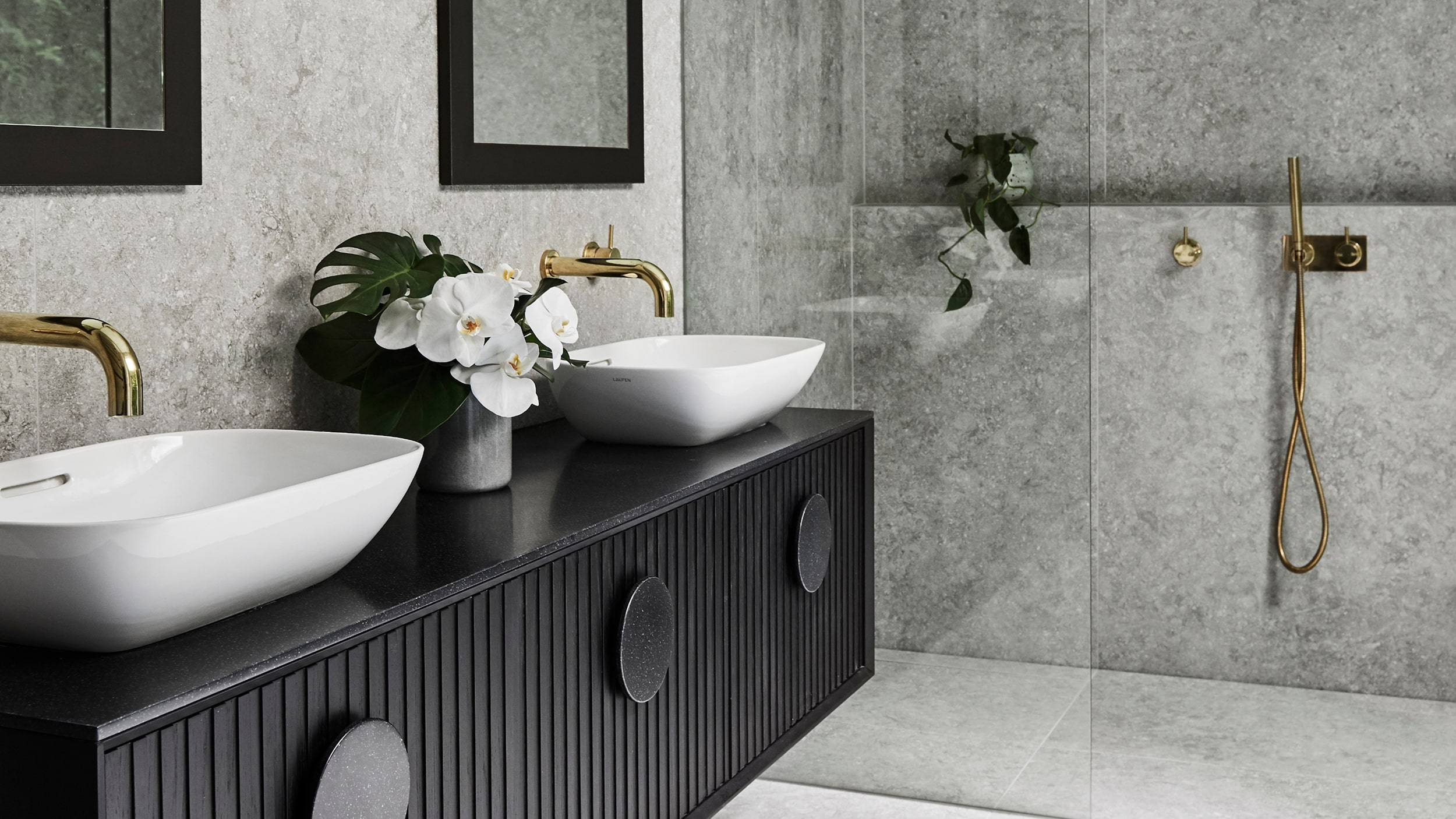 A striking bathroom with walk in shower, dark vanity, white basins and brushed gold tapware.