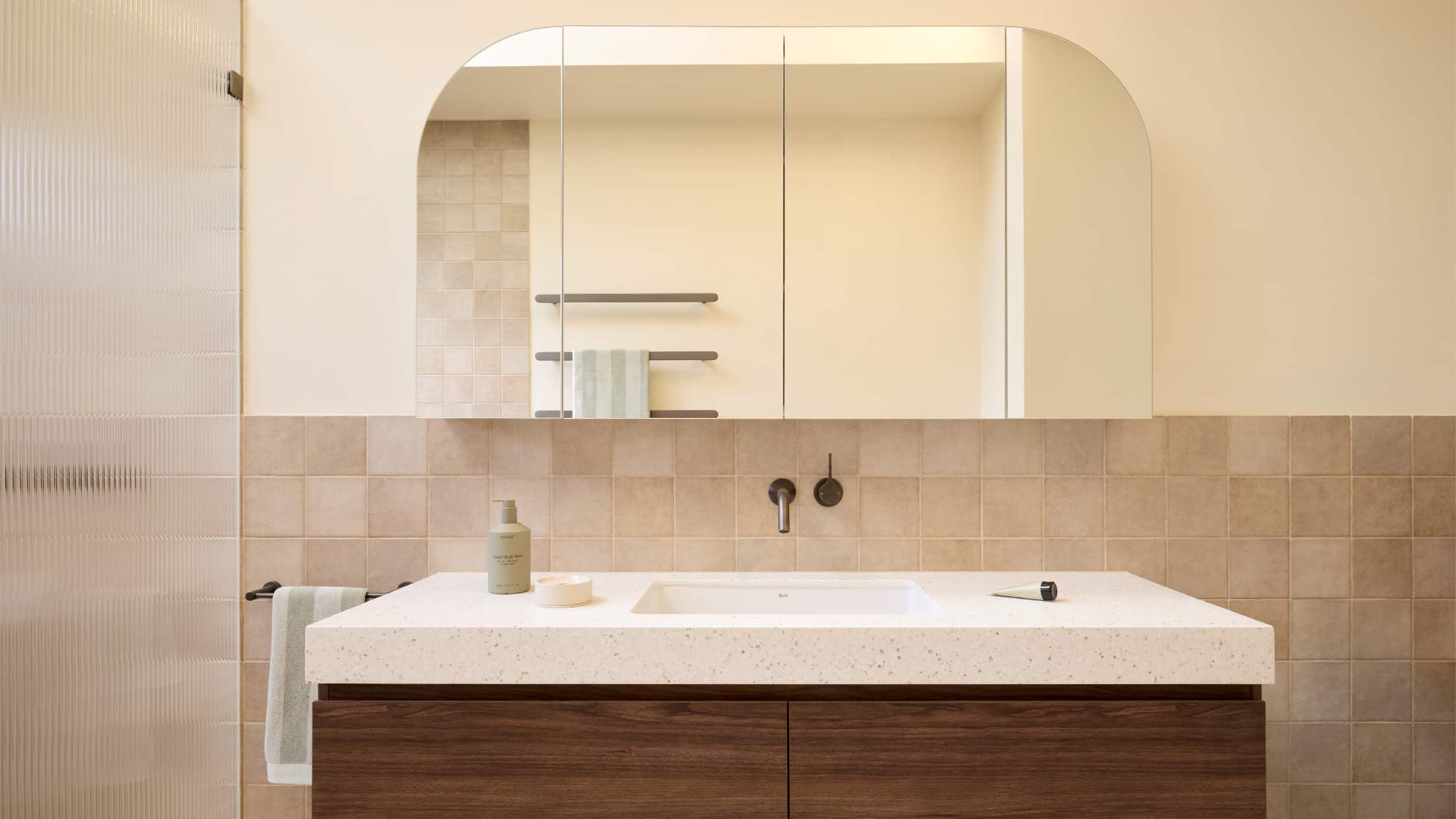 A spa-like bathroom featuring blush tile, deep walnut vanity, brushed gunmetal tapware, arched mirror cabinet