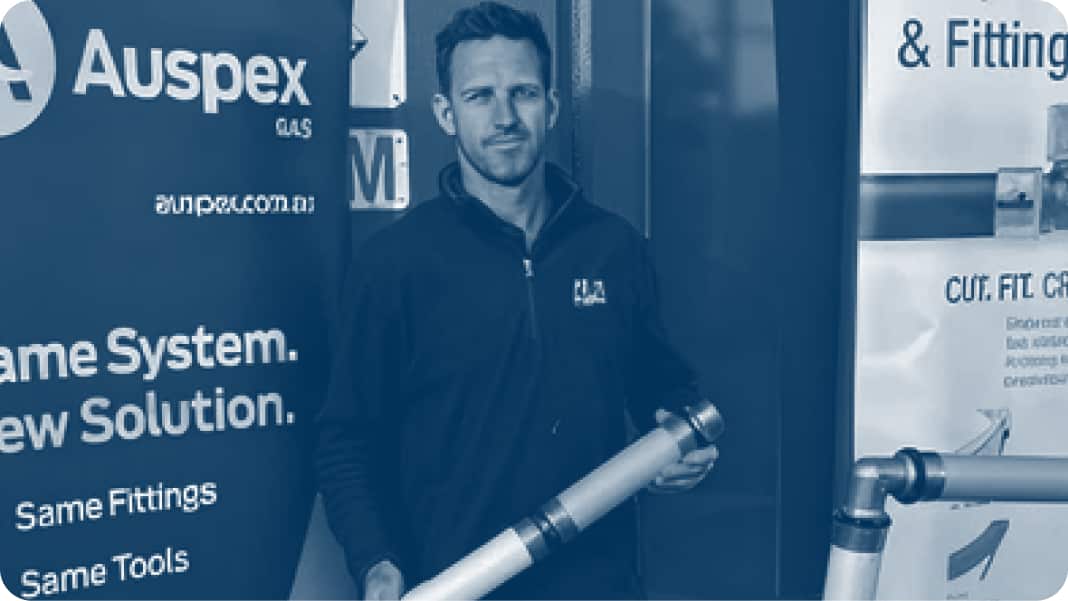 A plumber, standing in front of an Auspex banner, smiling at camera and holding a pipe.