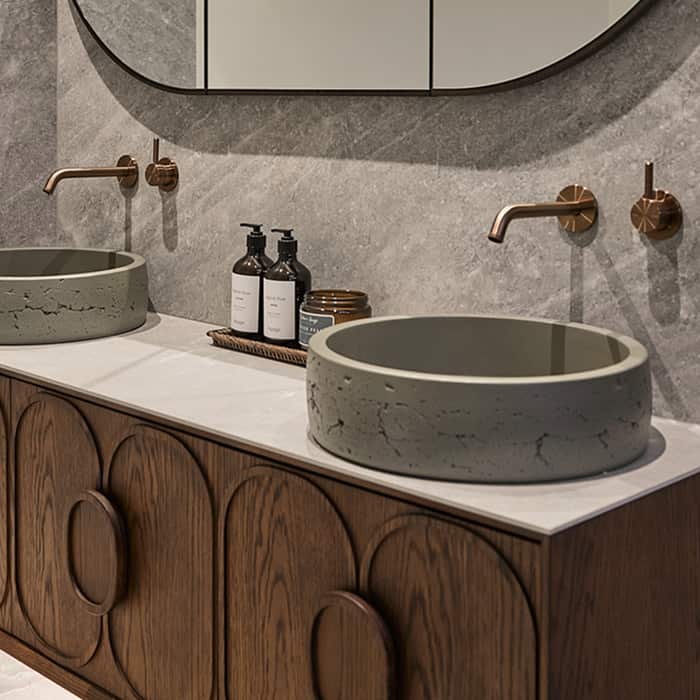 A timber vanity, two above counter basins, brushed bronze tapware, and oval mirror on a grey feature wall