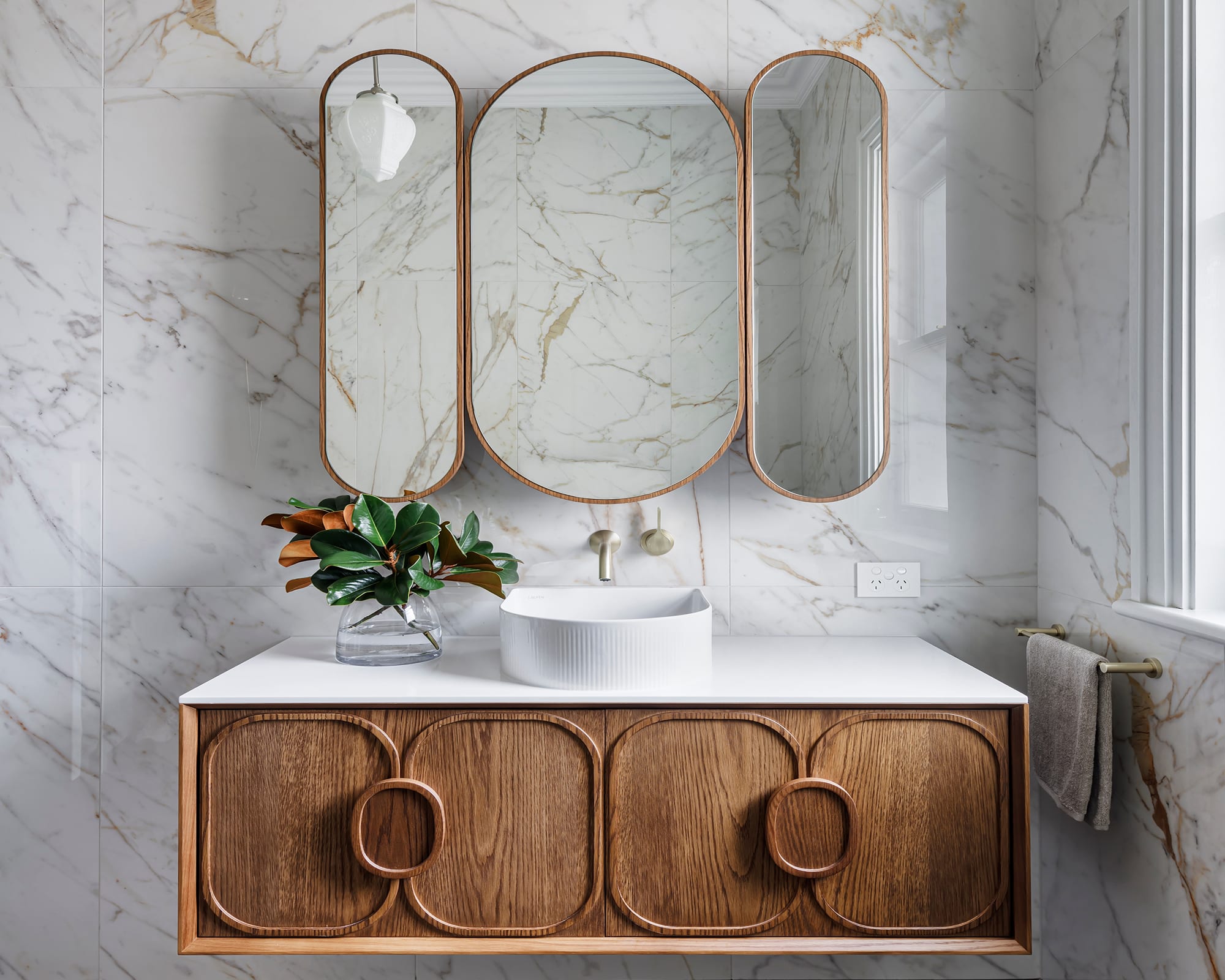 A classic, contemporary bathroom showcasing sleek marble walls paired with a warm wooden vanity, fluted basin and brushed gold tapware.