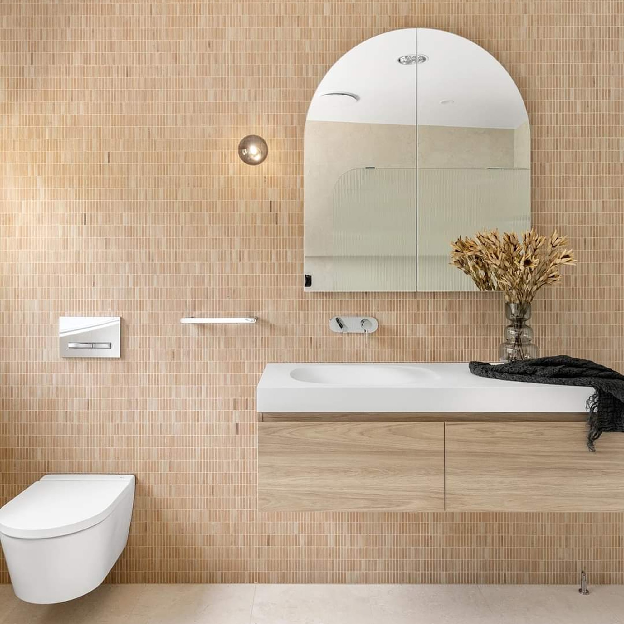 A modern bathroom featuring peach-coloured tiled walls, a wall hung toilet and a soft arched mirror and timber vanity.