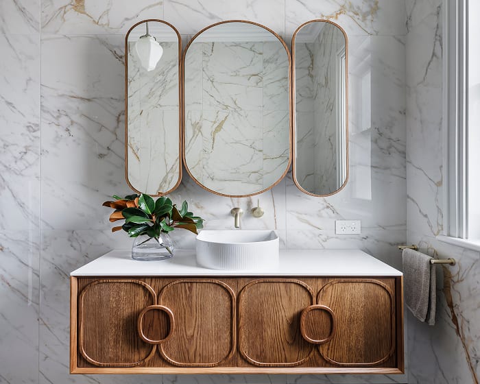 A classic, contemporary bathroom showcasing sleek marble walls paired with a warm wooden vanity, fluted basin and brushed gold tapware.