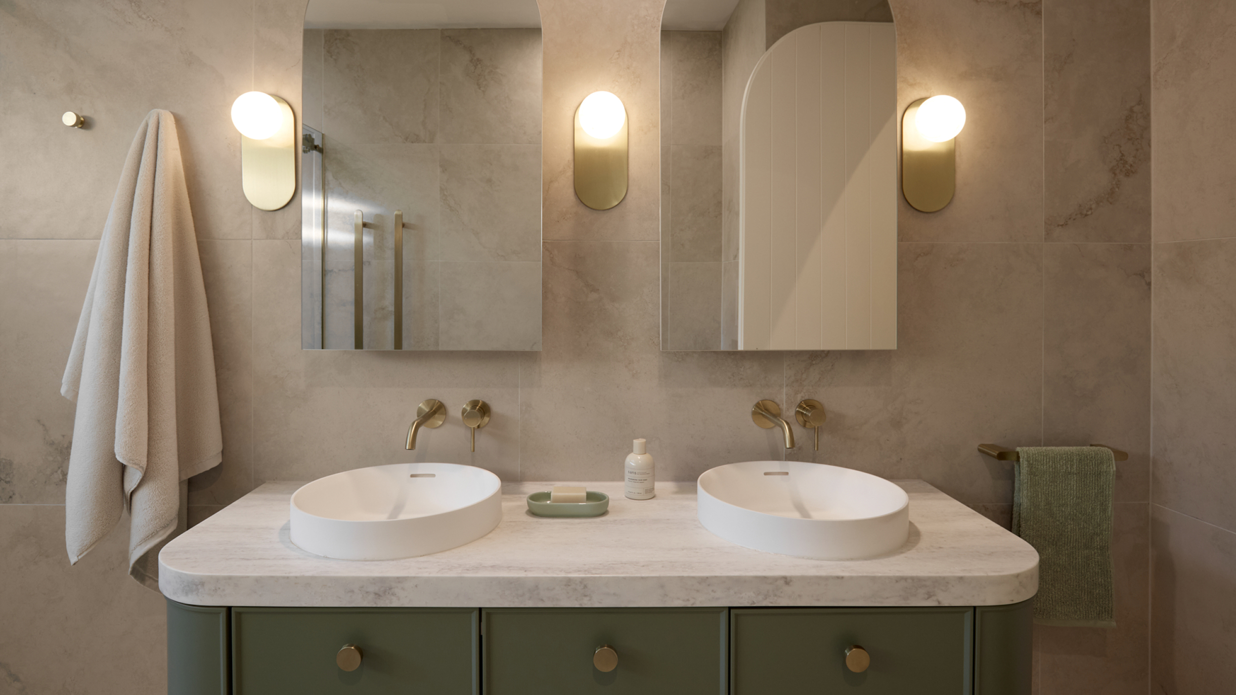 A spa-like bathroom featuring soft green vanity, two basins, two arched mirrors, framed by lights