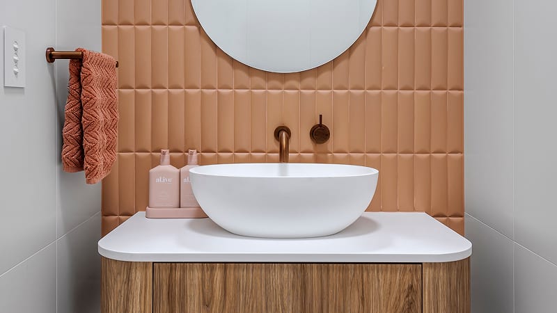 A modern bathroom basin with a round mirror above, peach-coloured tile backsplash, brown faucet, and pink soap dispensers on a wood vanity.