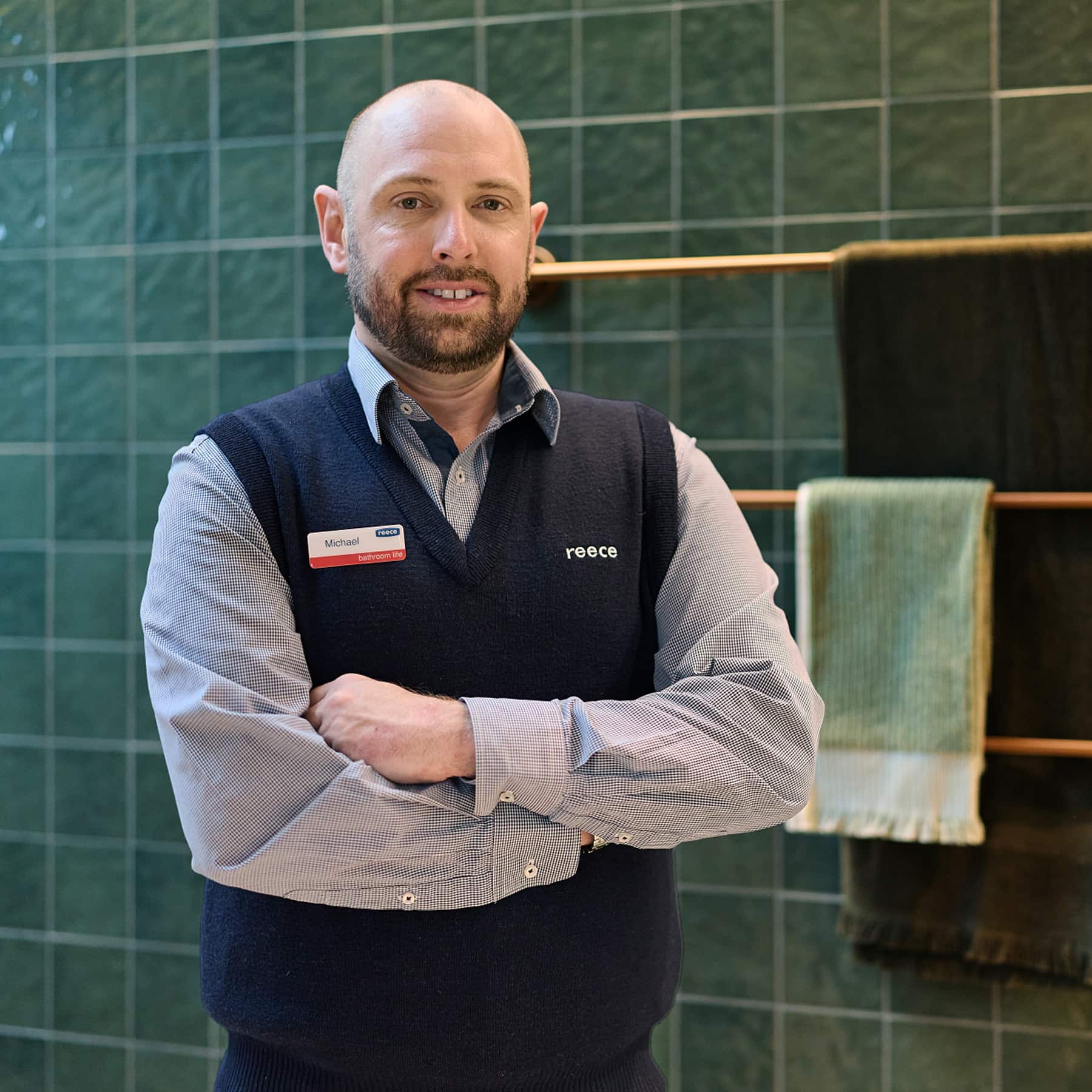 A man in a Reece uniform, standing in a green-tiled bathroom, smiling at the camera