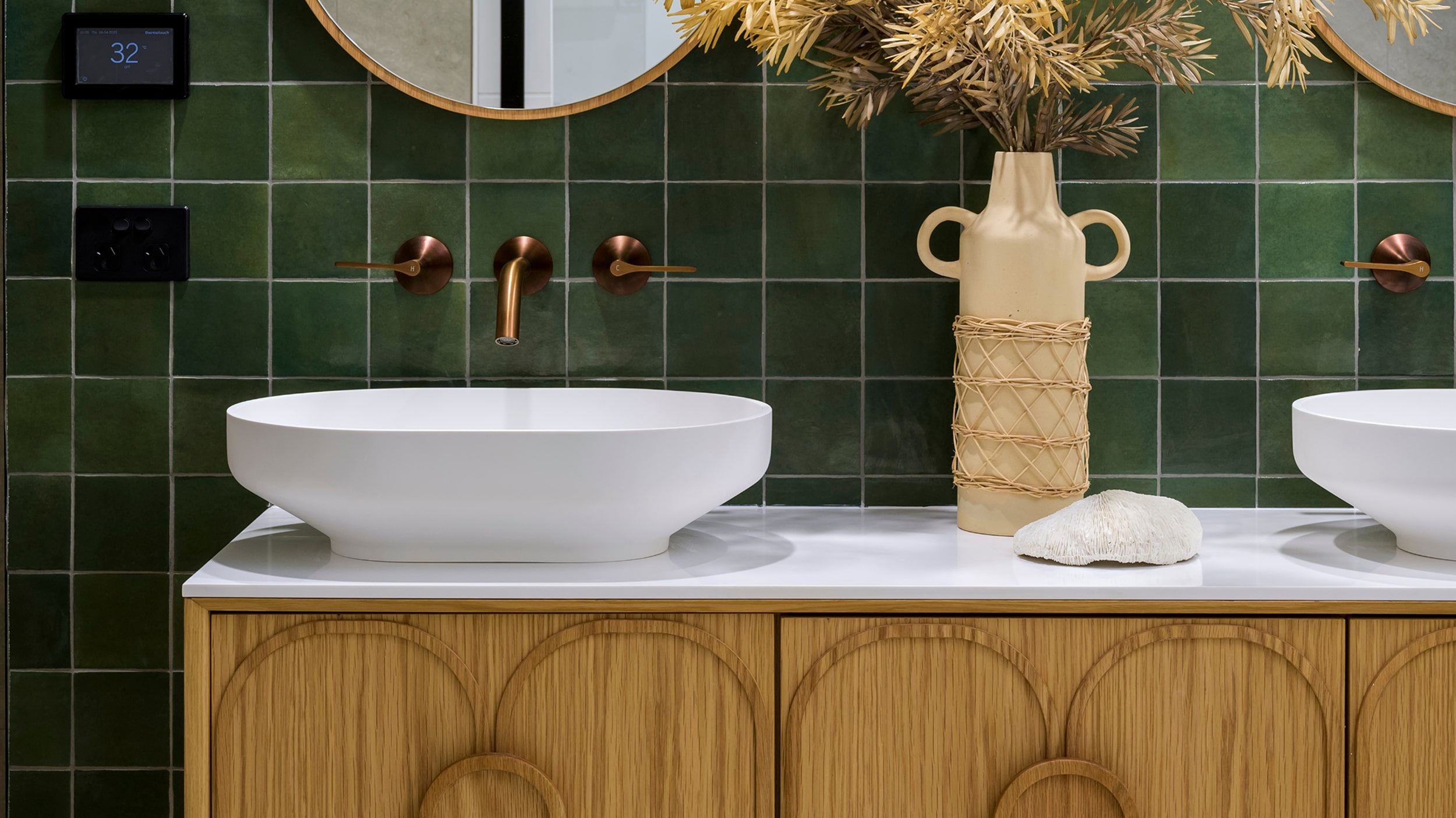 A modern bathroom featuring deep green tiles, a timber vanity, white basins and round mirrors.