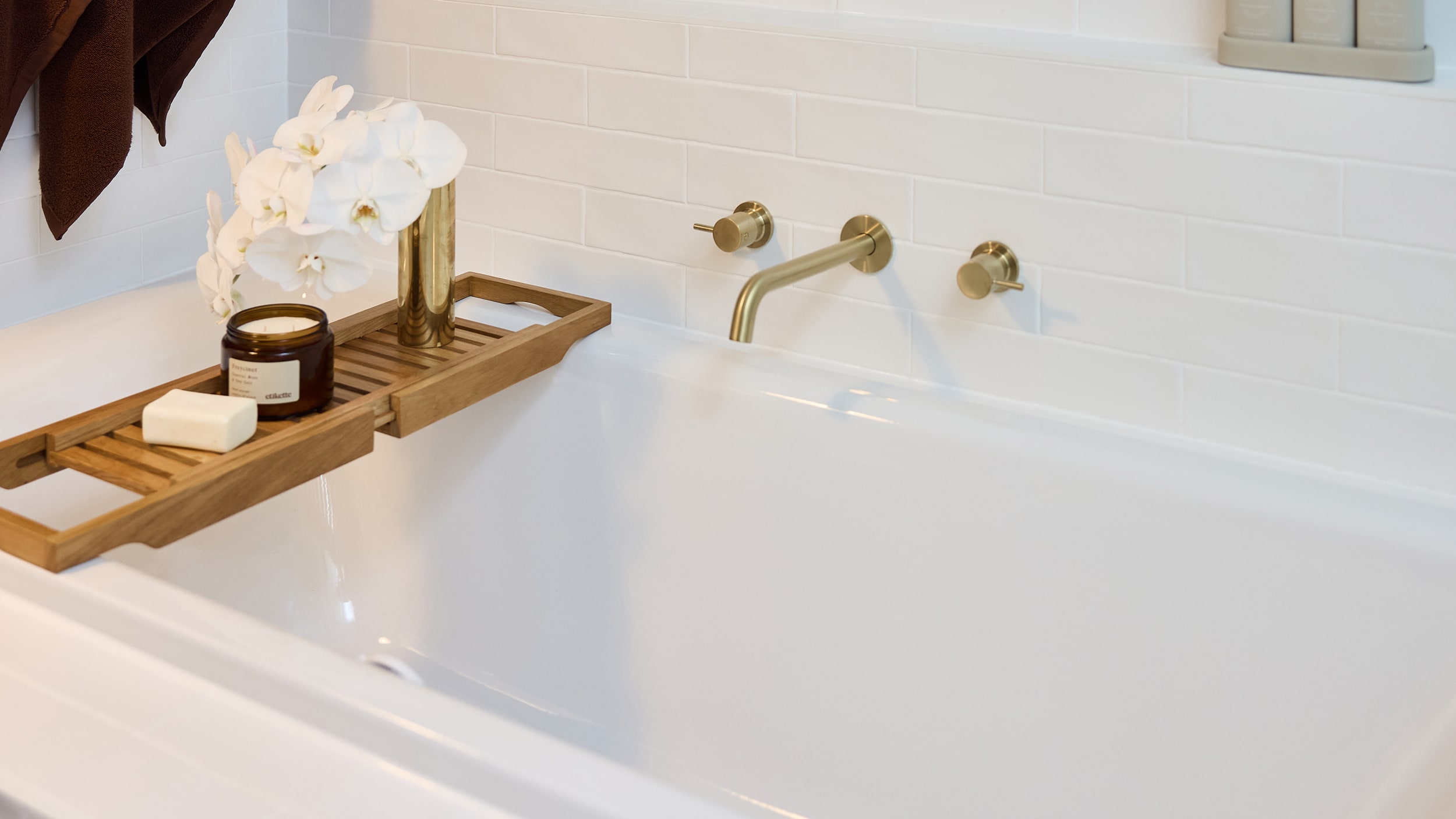 A close up of a white bathtub against a white, tiled wall with a wooden bath rack featuring flowers and a candle.