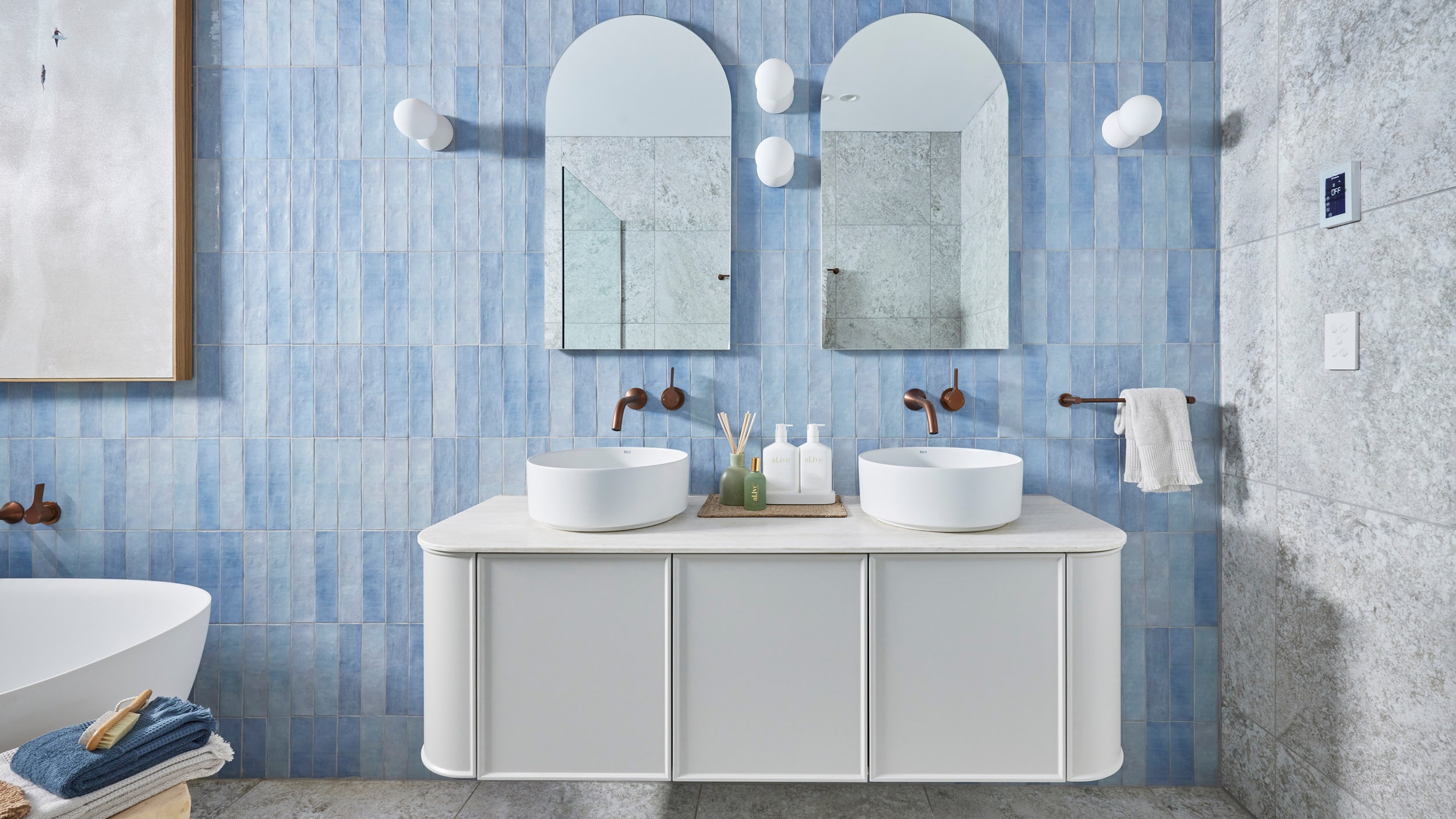 A light, bathroom featuring light blue tiled feature wall, white vanity and basins, arched mirrors and white deep bathtub.