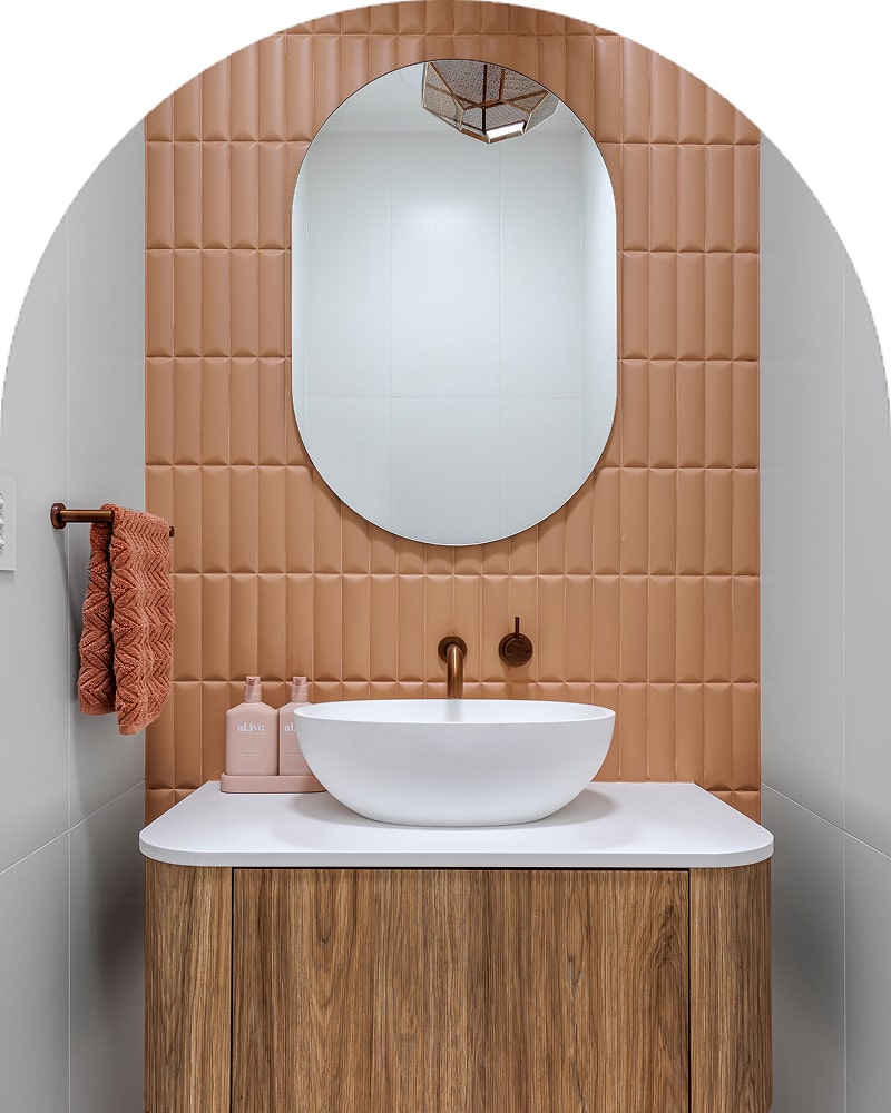 A small powder room featuring a white basin, timber vanity with a peach-tiled wall and oval mirror.
