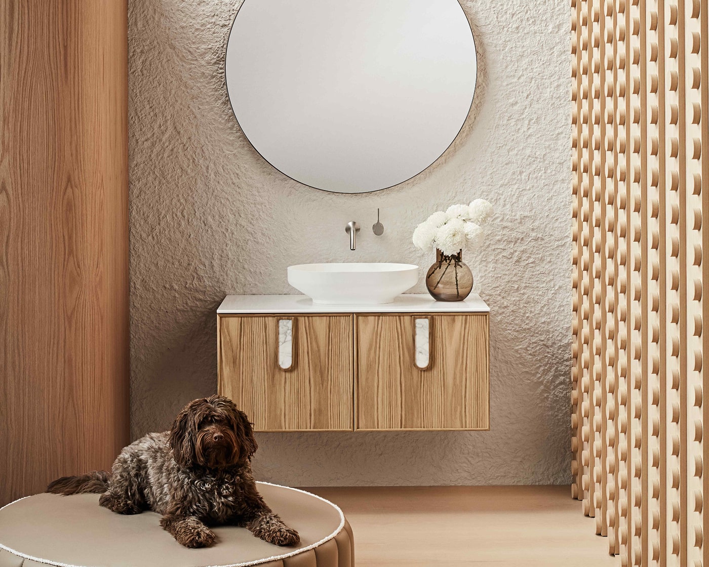 A sleek bathroom featuring timber vanity and feature walls, white basin, round mirror, and a dog.
