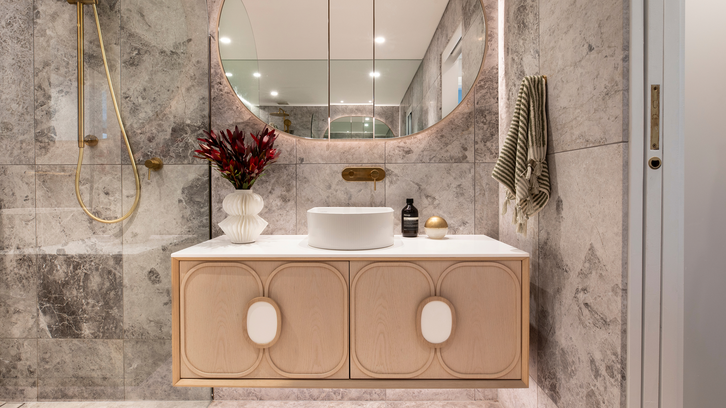 A stylish, bathroom featuring timber vanity, single basin, striking marble-tiled walls, and a backlit mirror.