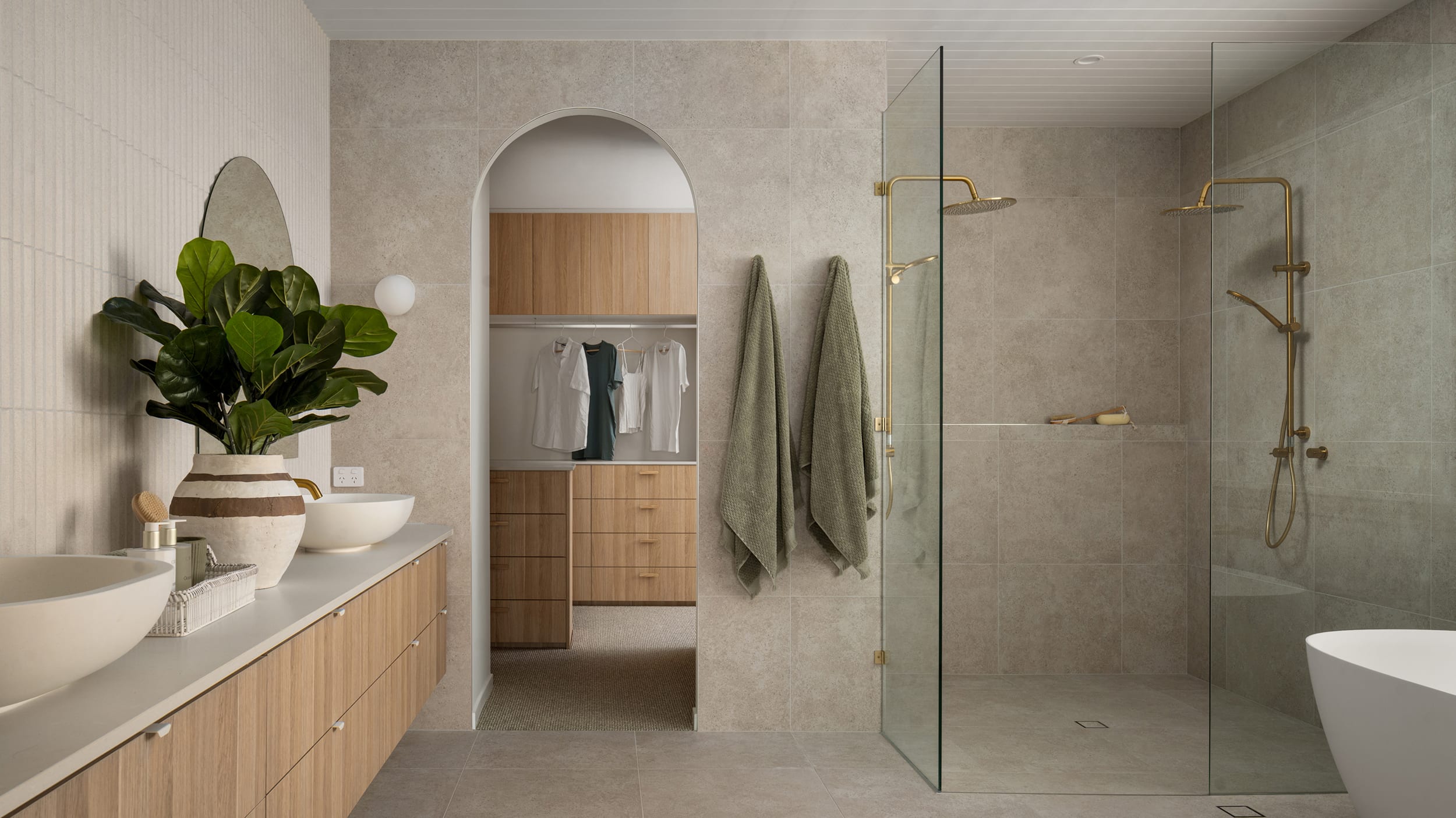 A spacious bathroom featuring a large walk-in shower, a natural, light wood vanity, styled with brushed gold tapware.