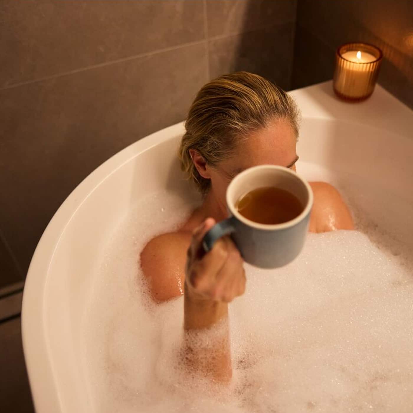 A woman relaxing in a bath in a darkened room lit with a candle, holding a cup of tea