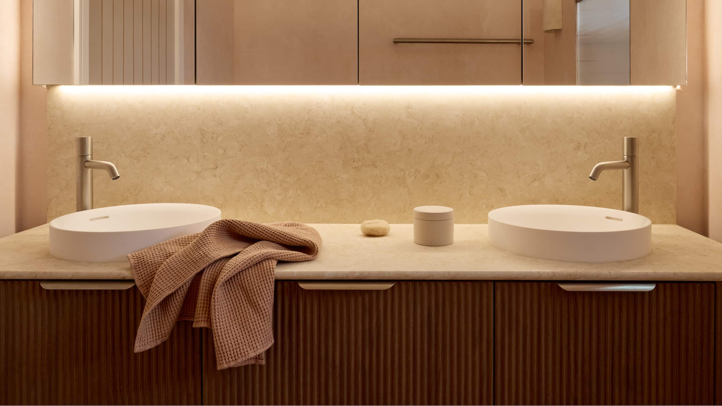 A timber bathroom vanity, two white above-counter basins with off-centred brushed nickel mixer taps, and a mirrored shaving cabinet with LED lighting