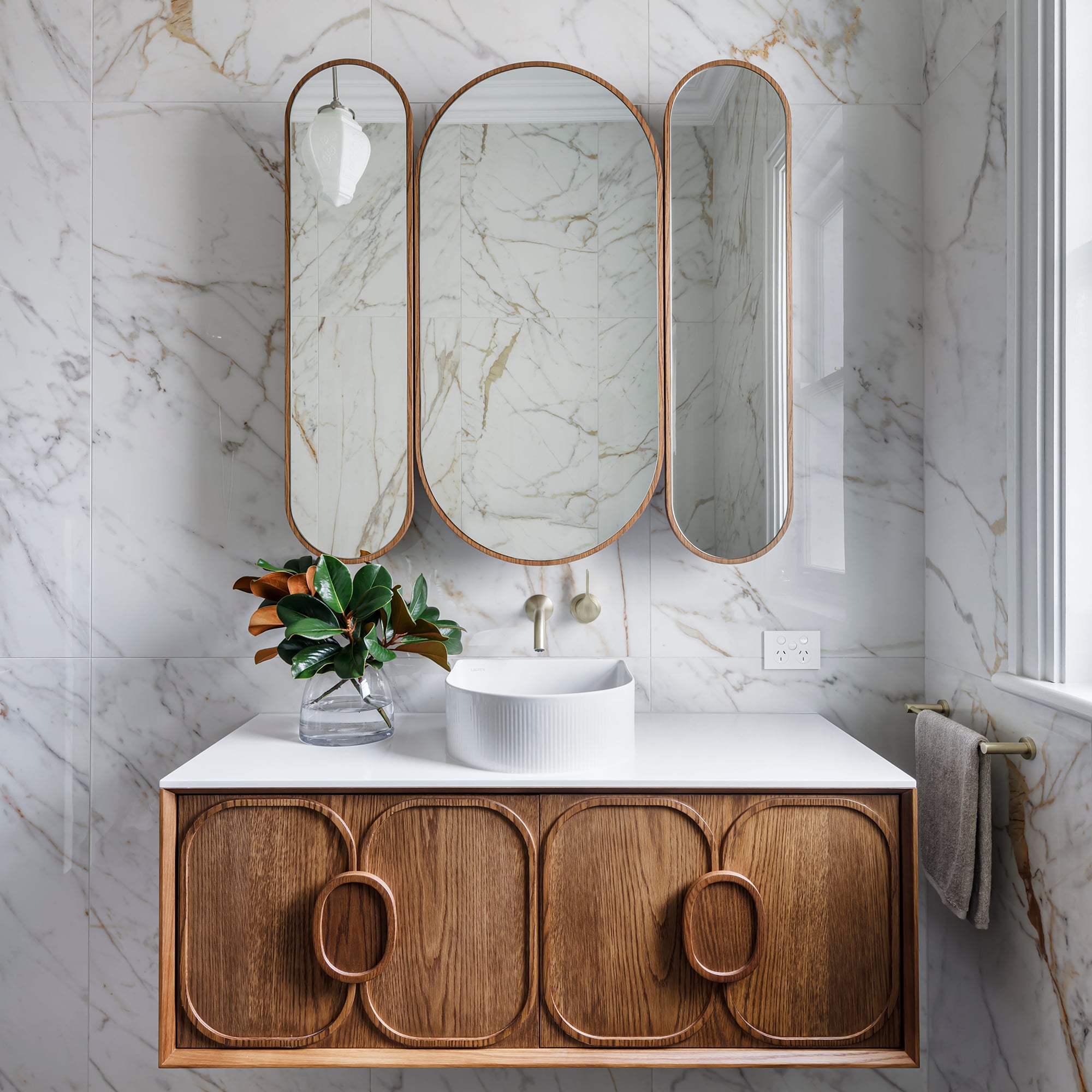 A classic, contemporary bathroom showcasing sleek marble walls paired with a warm wooden vanity, fluted basin and brushed gold tapware.