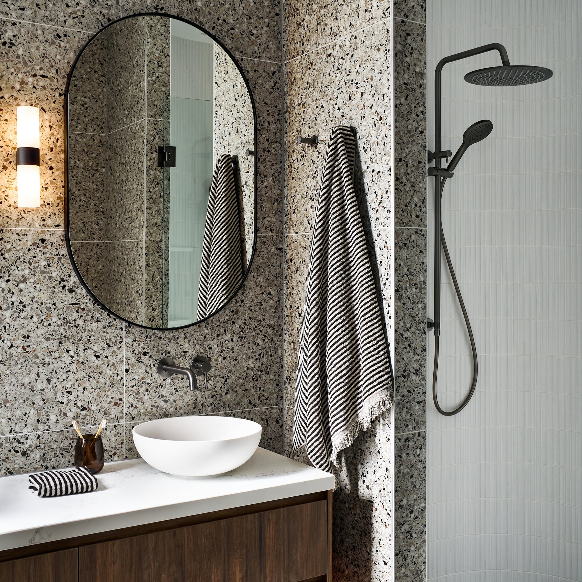 An elegant, moody bathroom with a white basin paired with a round mirror, dark metal tapware and a dark wooden vanity, styled with striped towels.