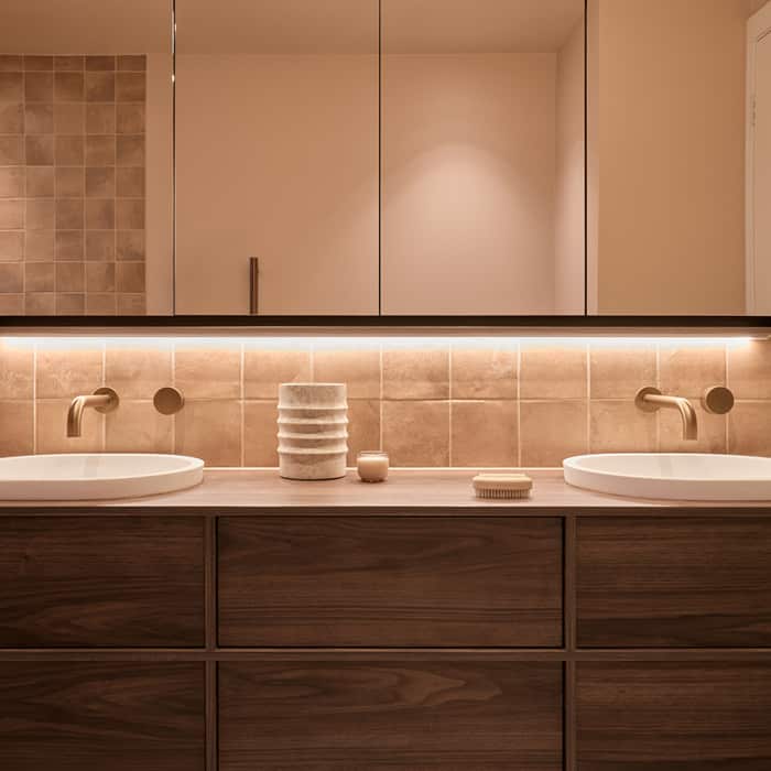 A spa-like bathroom featuring muted tile, timber vanity with two basins, mirrored cabinet with LED lighting, and brushed bronze tapware