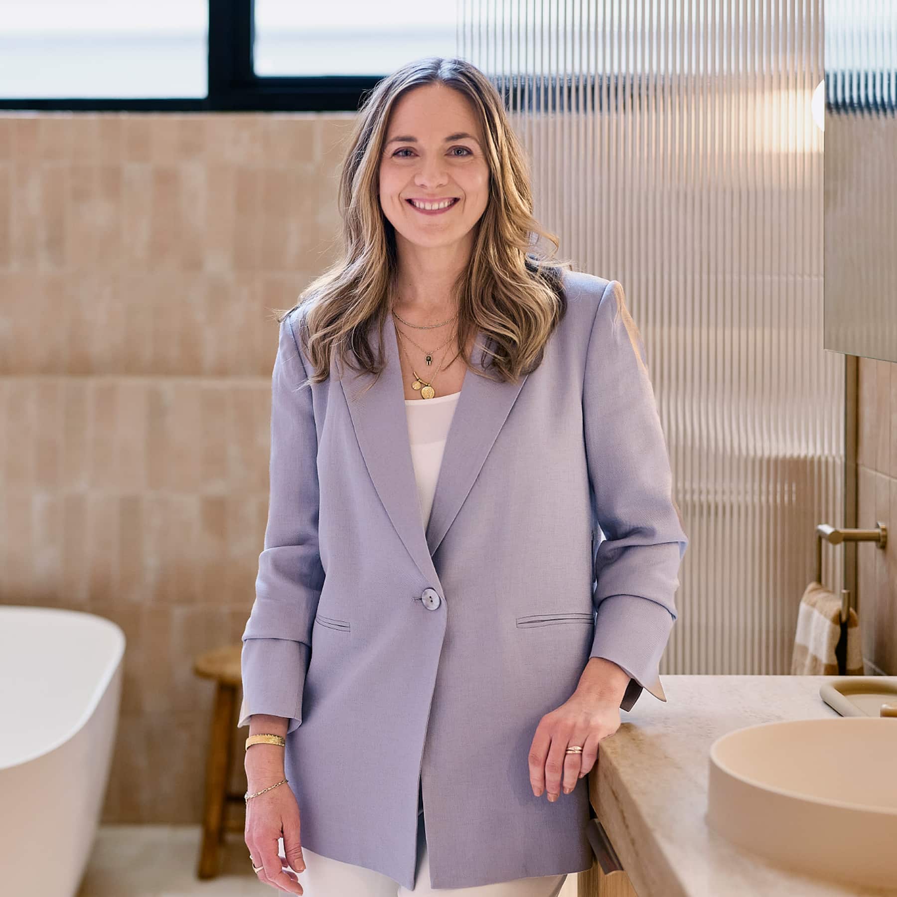 A professional looking woman in a light purple blazer, standing in a spa-like bathroom and smiling directly at the camera.