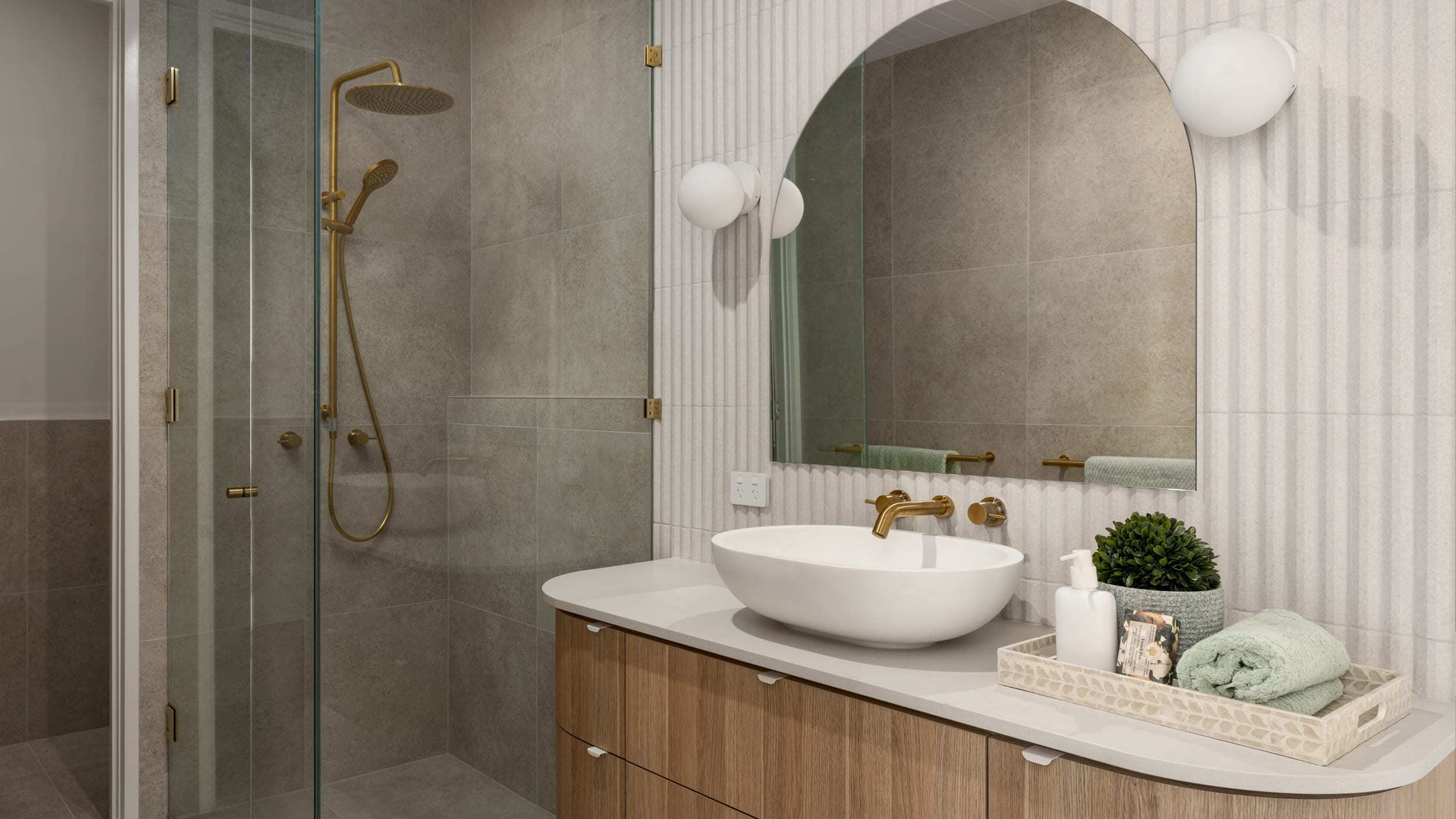 A stylish, clean bathroom featuring a white basin, light wood vanity, gold tapware and a natural, light wood vanity.