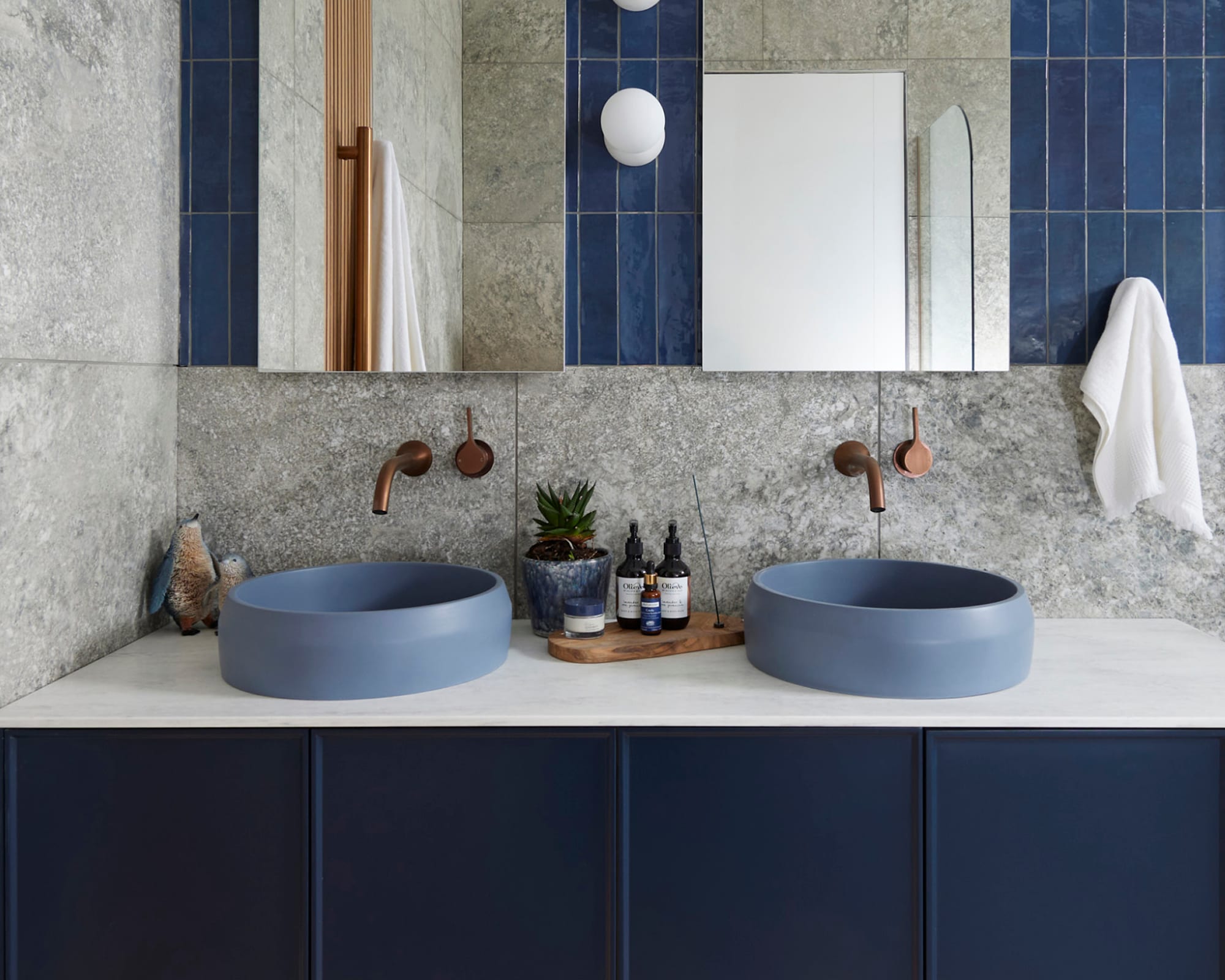 Modern bathroom with two round blue basins, wall-mounted faucets, a wooden tray with plants and toiletries, and a towel hanging on the right.