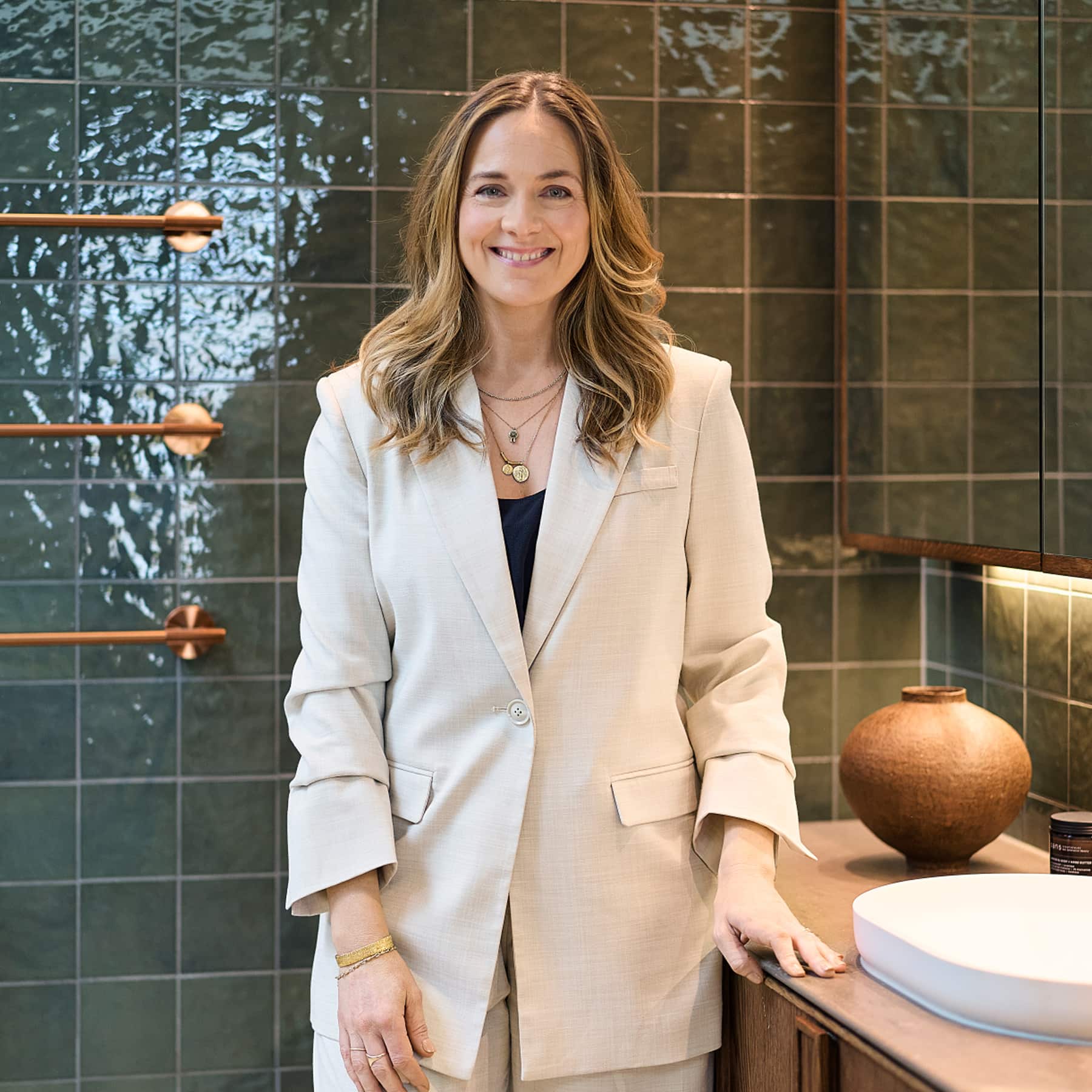 A smartly-dressed woman, standing in a green-tiled bathroom, smiling at the camera