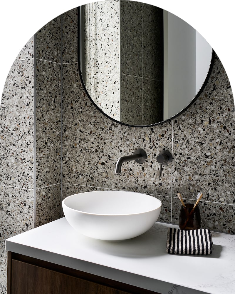 An elegant, moody bathroom with a white basin paired with a round mirror, dark metal tapware and a dark wooden vanity.