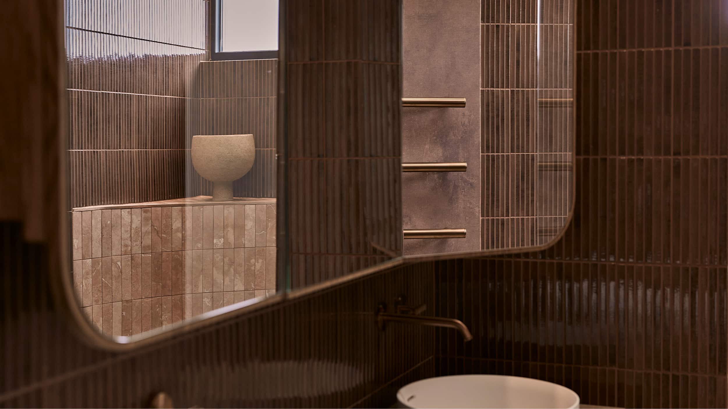 A mirrored shaving cabinet, reflecting a moody bathroom, featuring berry-hued tile and brushed bronze fittings