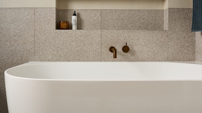 A sleek, back-to-wall style bathtub paired with dark metal tapware, in a bathroom with a recessed shelf above it displaying toiletries.